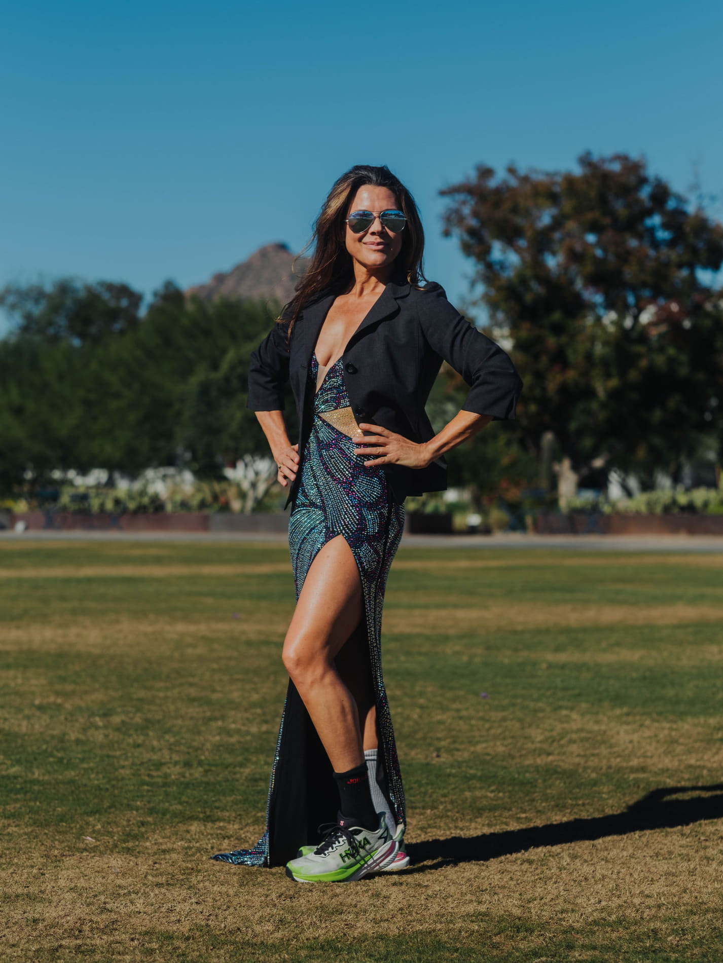 A woman standing outdoors on a sunny day in a blazer and sequined dress, with trees and a mountain behind her.