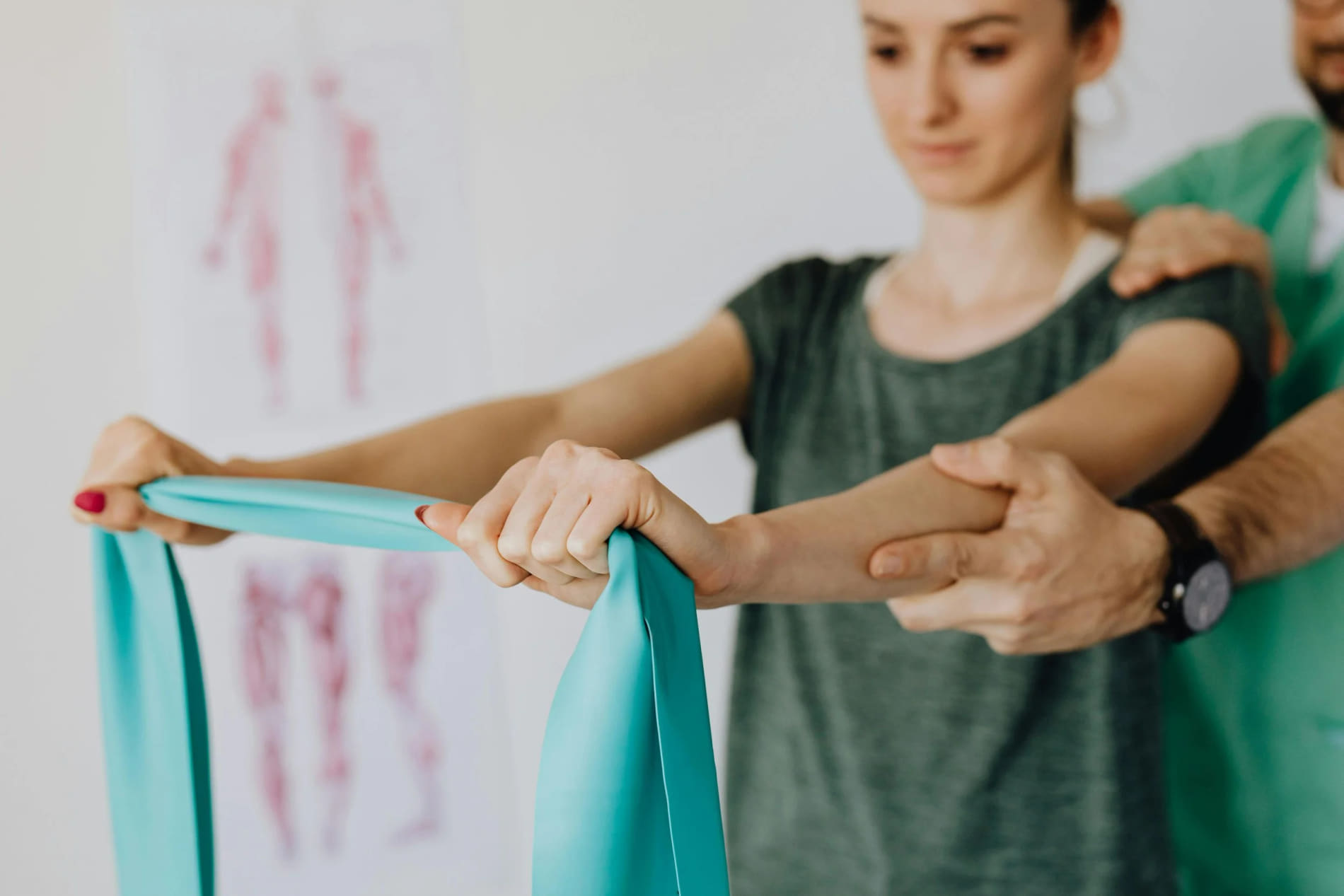 A woman in a grey shirt receiving physical therapy on her arm from a therapist, with a diagram of the human body in the background.