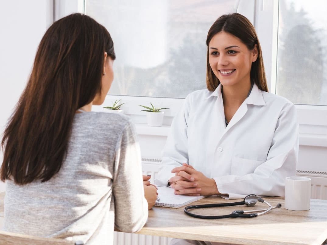 A doctor and a patient having a consultation in a medical office, with a stethoscope on the table and plants on the window sill.
