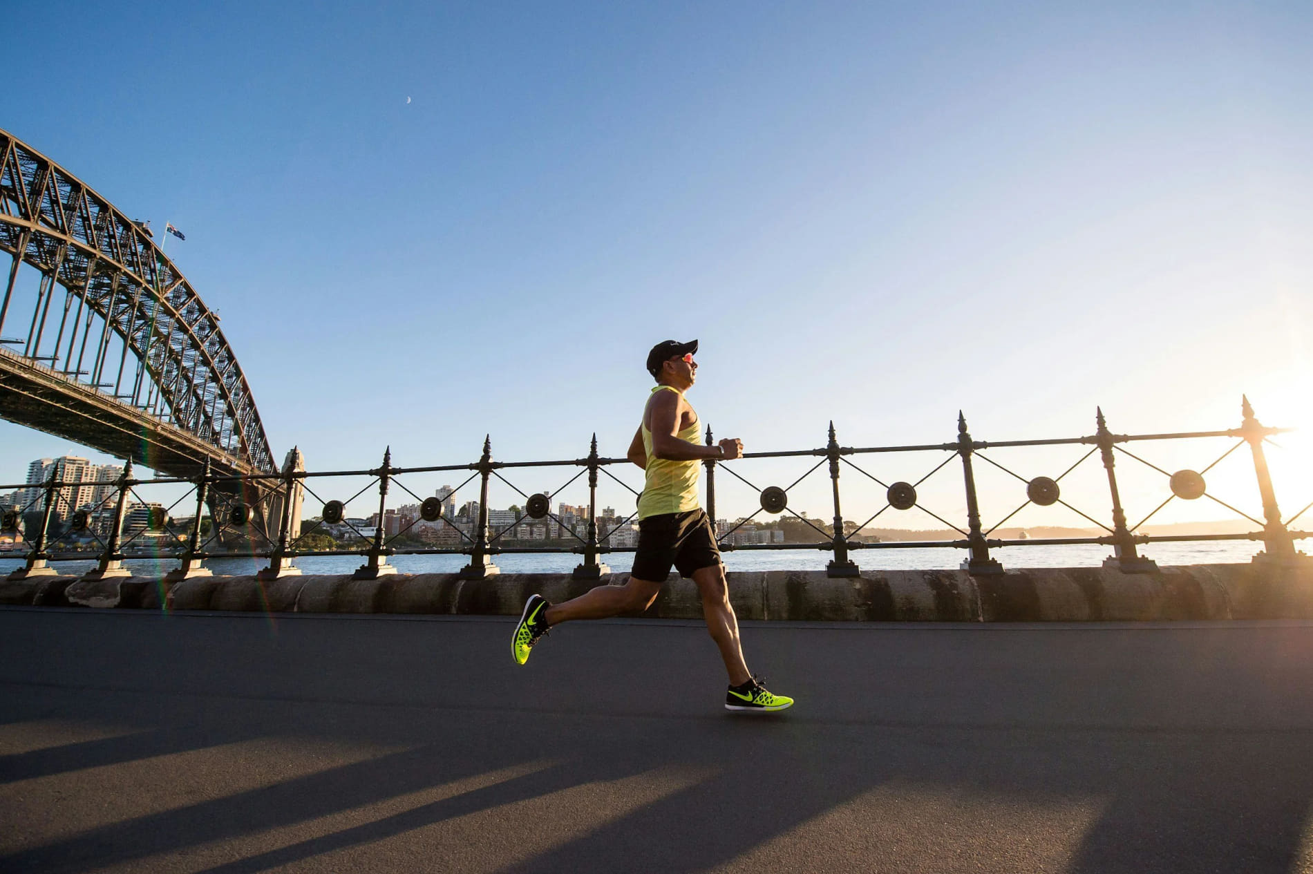 A man running along a waterfront road during sunset, with a bridge and city skyline in the background.