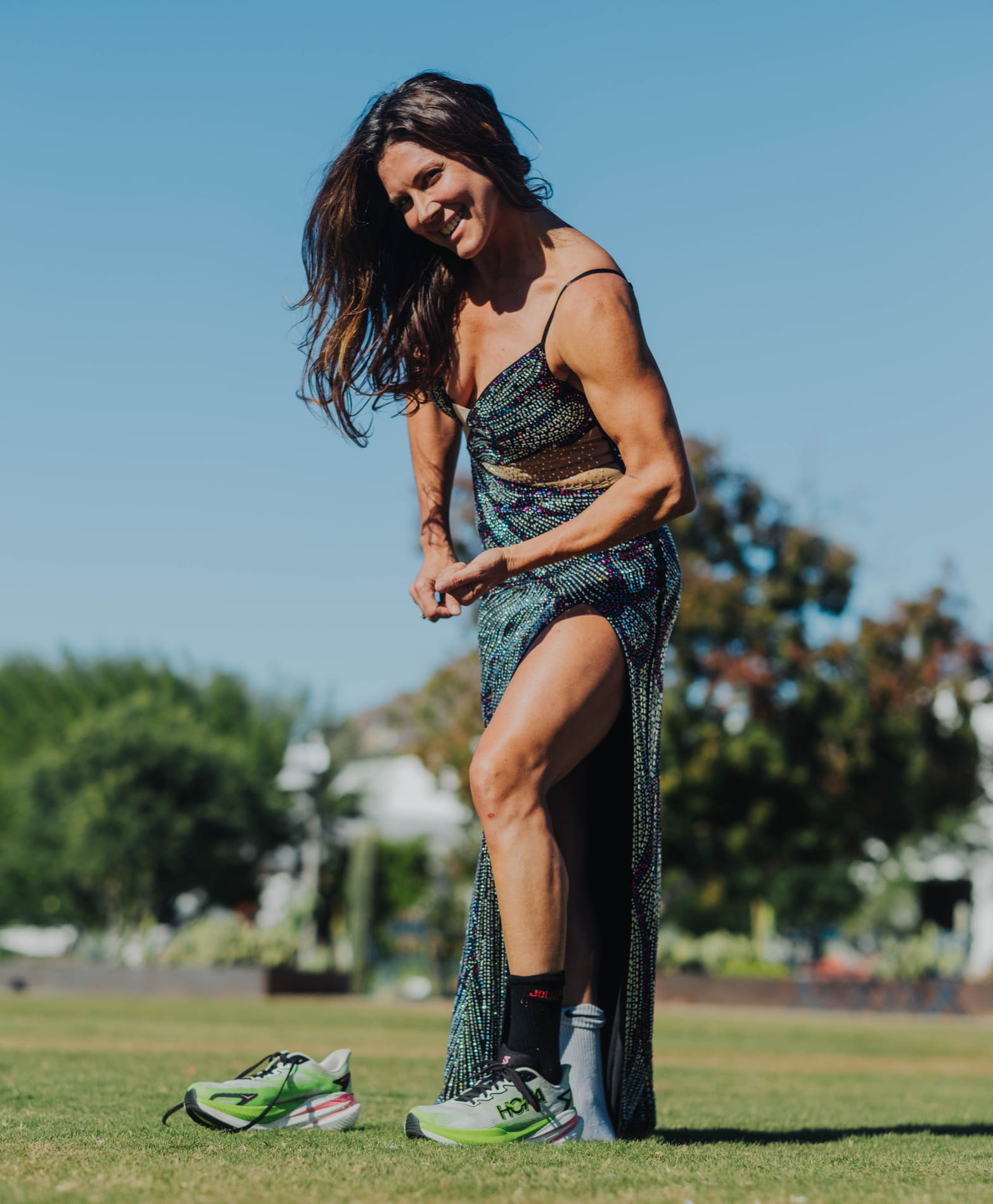 A woman adjusting her running shoes on a grassy field under a clear blue sky.